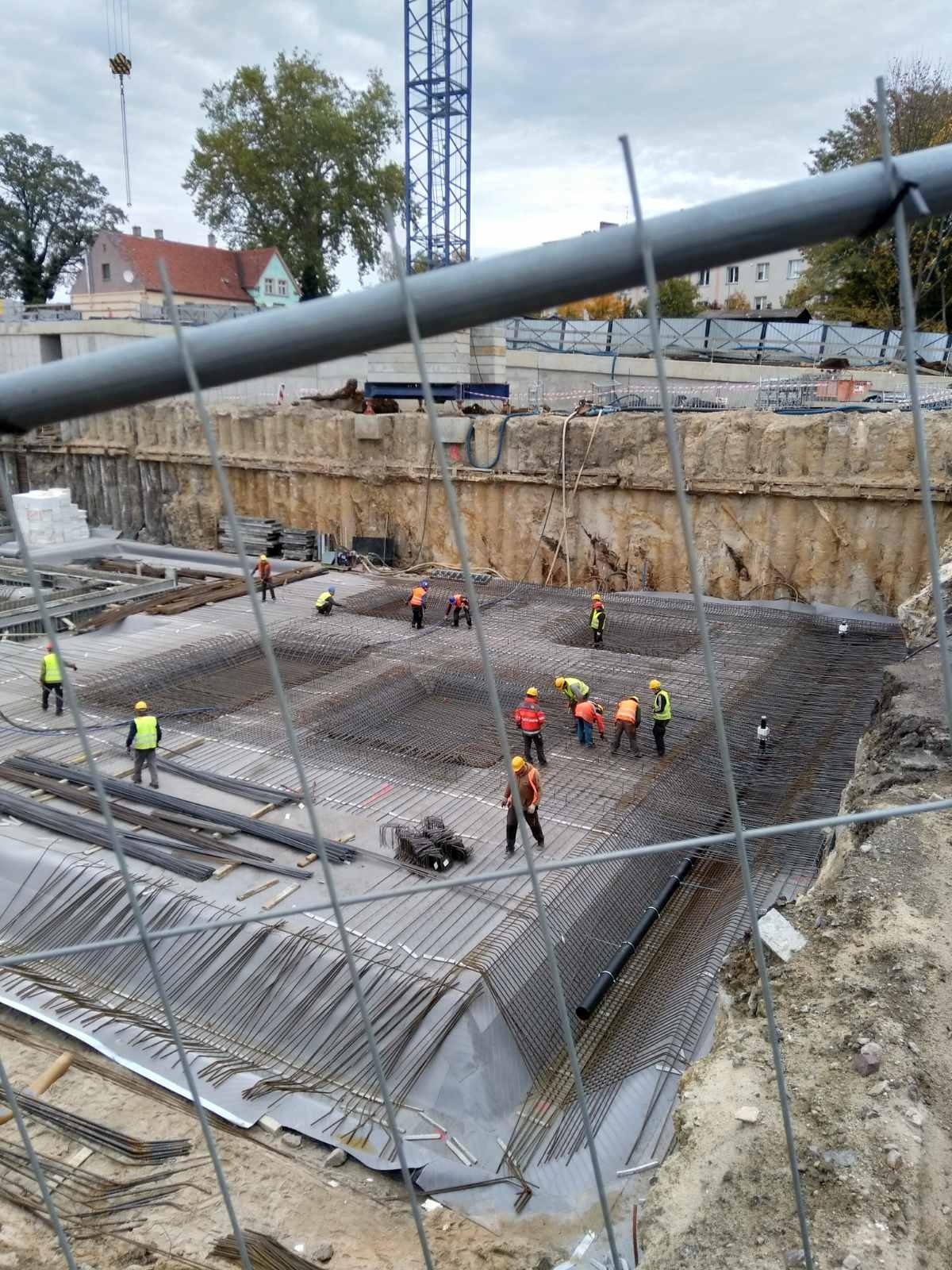 Prace zbrojarskie na placu budowy, widok przez ogrodzenie; robotnicy w kaskach i kamizelkach układają siatkę z prętów na folii, w tle wykop i żuraw budowlany.