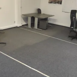 Profesjonalne urządzenie do ekstrakcyjnego czyszczenia dywanów z niebieskim wężem w biurze z szarym dywanem, widoczne biurka i krzesła.