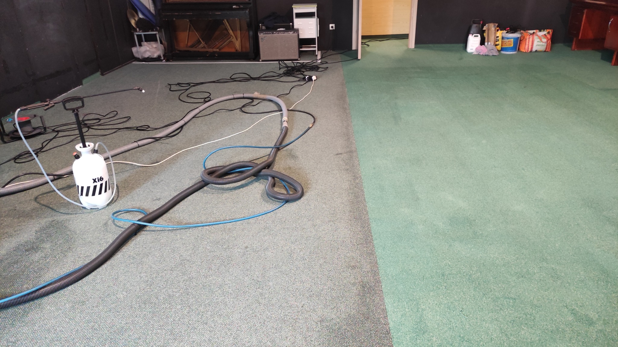 Fragment zielonej wykładziny dywanowej, z widoczną różnicą w odcieniu po lewej stronie, gdzie leżą węże i spryskiwacz do czyszczenia, sugerujący etap czyszczenia.