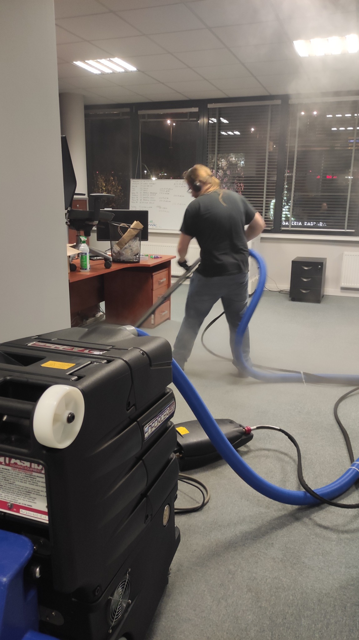 Pracownik w trakcie czyszczenia wykładziny biurowej za pomocą profesjonalnego ekstraktora do czyszczenia dywanów, widoczny wylot pary i niebieski wąż ssący.