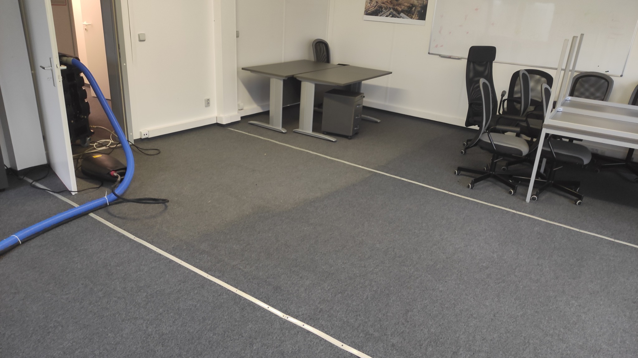 Profesjonalne urządzenie do ekstrakcyjnego czyszczenia dywanów z niebieskim wężem w biurze z szarym dywanem, widoczne biurka i krzesła.
