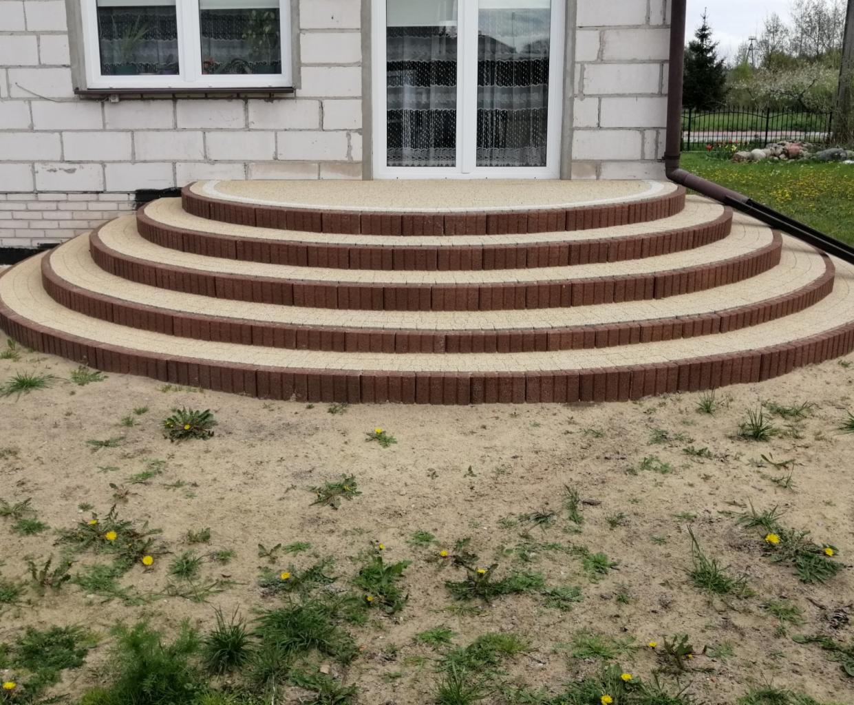 Półokrągłe schody wejściowe z kostki brukowej w dwóch kolorach, prowadzące do drzwi wejściowych, widoczne chwasty na piasku przed schodami.