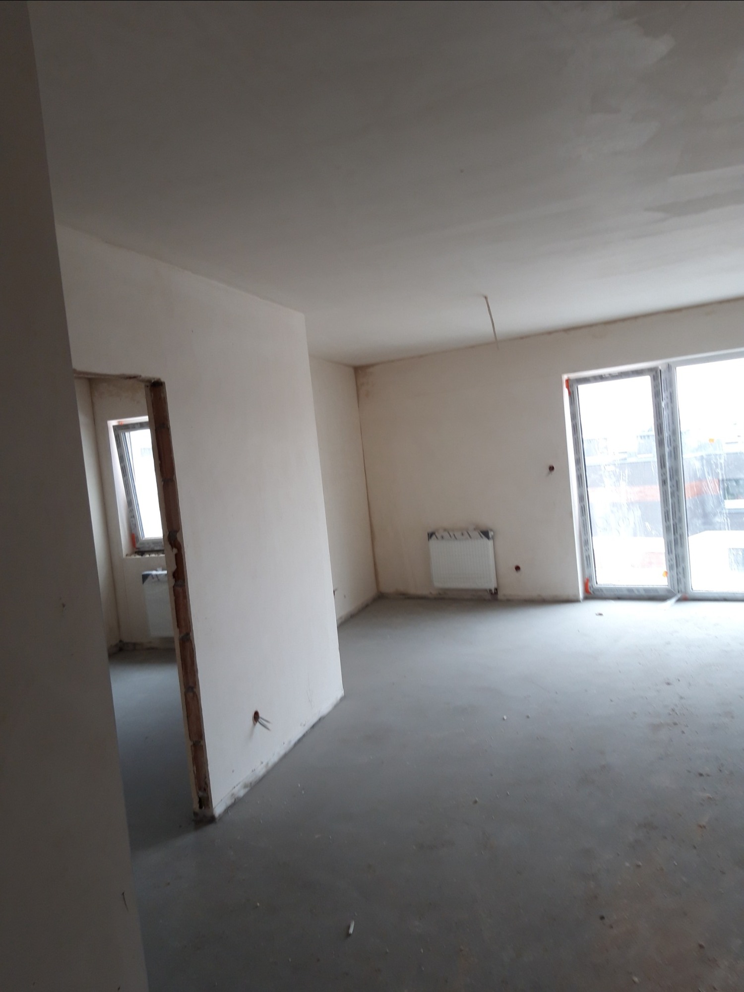 Surowe wnętrze mieszkania w trakcie wykańczania z widocznymi ścianami po nałożeniu gładzi, przygotowanymi do dalszego etapu prac, z oknami i grzejnikami.