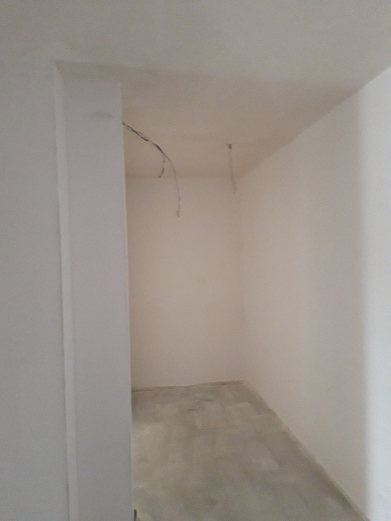 Wnętrze pomieszczenia w trakcie prac wykończeniowych, ściany pomalowane na biało, widoczne wystające kable z sufitu, podłoga w stanie surowym.
