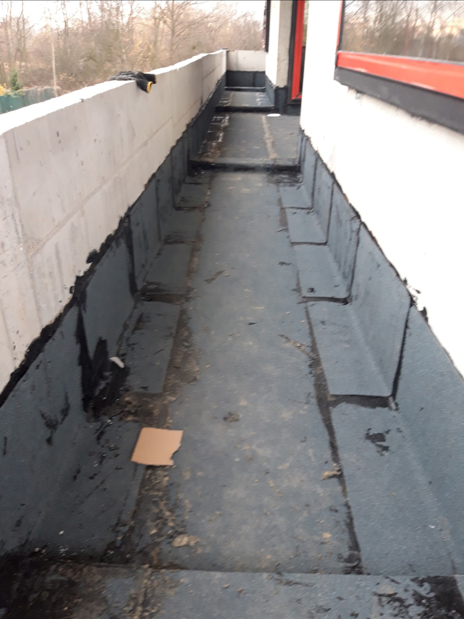 Hydroizolacja balkonu w trakcie prac, widoczne fragmenty papy bitumicznej i ślady smoły, surowe ściany i fragment kartonu na podłożu.