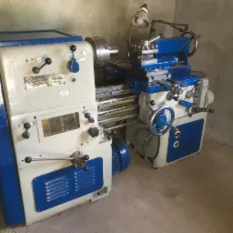 Maszyna tokarska w warsztacie, malowana na biało i niebiesko, z widoczną lampką oświetlającą obszar roboczy i licznymi dźwigniami sterującymi.