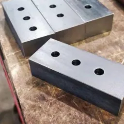 Precyzyjnie obrobione, metalowe prostopadłościany z trzema otworami każdy, ułożone na blacie roboczym. Widoczne ślady obróbki.