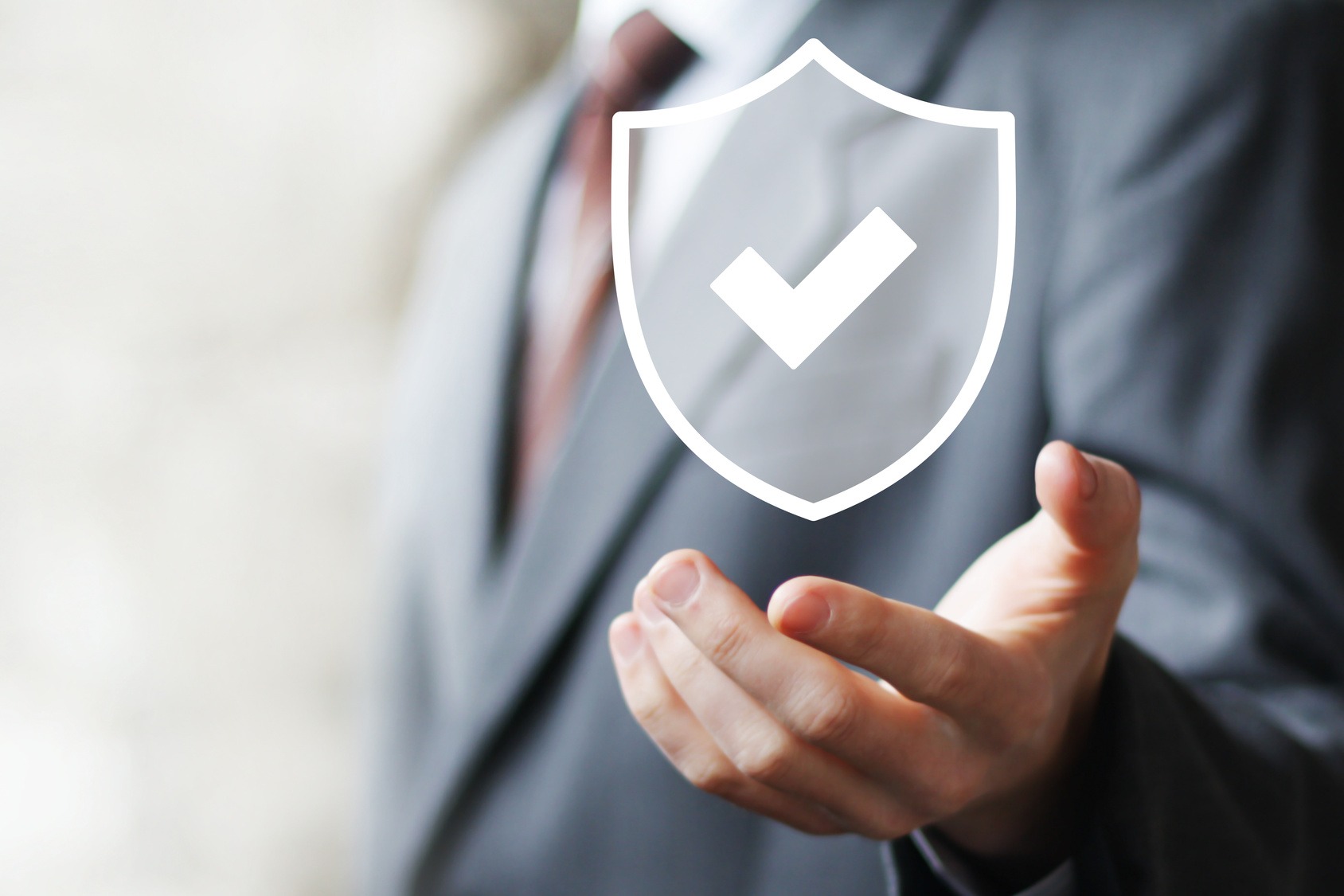 Mężczyzna w garniturze prezentuje na otwartej dłoni białą ikonę tarczy z symbolem akceptacji, sugerującą bezpieczeństwo i ochronę danych.