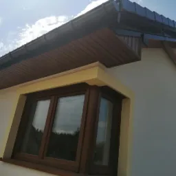 Fragment elewacji domu jednorodzinnego z drewnianą podbitką dachową, brązowymi oknami i żółtymi obramowaniami okiennymi, widziany pod kątem w słoneczny dzień.