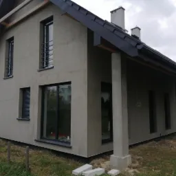 Dom jednorodzinny z szarą, surową elewacją w trakcie ocieplania, widoczne okna z czarnymi ramami, fragment dachu z kominami i elementy drewniane konstrukcji dachu.