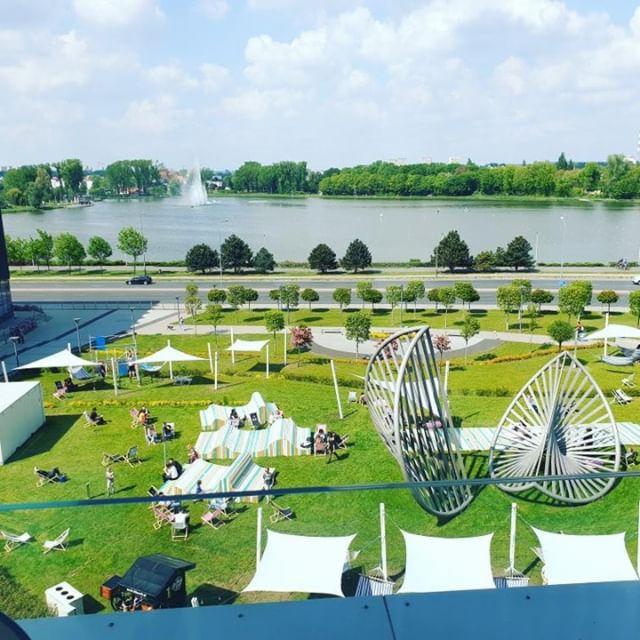 Widok z góry na park miejski z jeziorem, fontanną i leżakami. Na pierwszym planie metalowe rzeźby i białe zadaszenia.