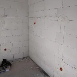 Surowe ściany z betonu komórkowego z wyfrezowanymi bruzdami i puszkami pod instalację elektryczną, widoczne przewody i rury osłonowe na podłodze.