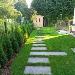 Zadbany ogród z równo przystrzyżonym trawnikiem, ścieżką z płyt betonowych prowadzącą do drewnianej altany, rządkiem tuj przy ogrodzeniu i wiszącym fotelem na tle zieleni.