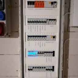 Rozdzielnica elektryczna z zainstalowanymi zabezpieczeniami, wyłącznikami różnicowoprądowymi, licznikiem energii elektrycznej ORNO i opisami obwodów, zamontowana na ścianie z surowego betonu.