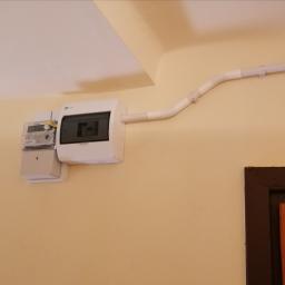 Elstart usługi elektryczne Michał Bejger - Zamontowany licznik elektryczny z białą skrzynką bezpiecznikową i peszlem na beżowej ścianie, widoczny fragment brązowej futryny drzwi.