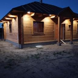 Elstart usługi elektryczne Michał Bejger - Drewniany dom jednorodzinny oświetlony zewnętrznymi lampami punktowymi pod okapem dachu, widok wieczorny z ziemistym podłożem.