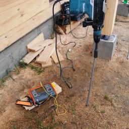 Elstart usługi elektryczne Michał Bejger - Młot udarowy Makita wbija uziom; obok leży miernik elektryczny z przewodami, na tle drewnianej konstrukcji w surowym stanie.