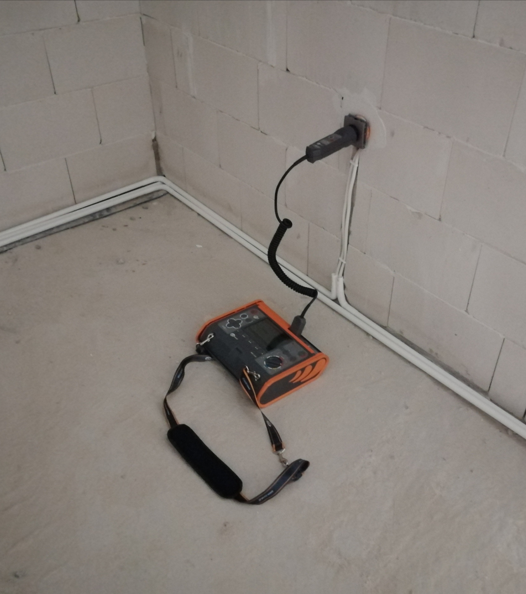 Miernik elektryczny z podłączoną sondą w gnieździe, widoczne kable elektryczne w listwach przypodłogowych i ściana z betonu komórkowego.