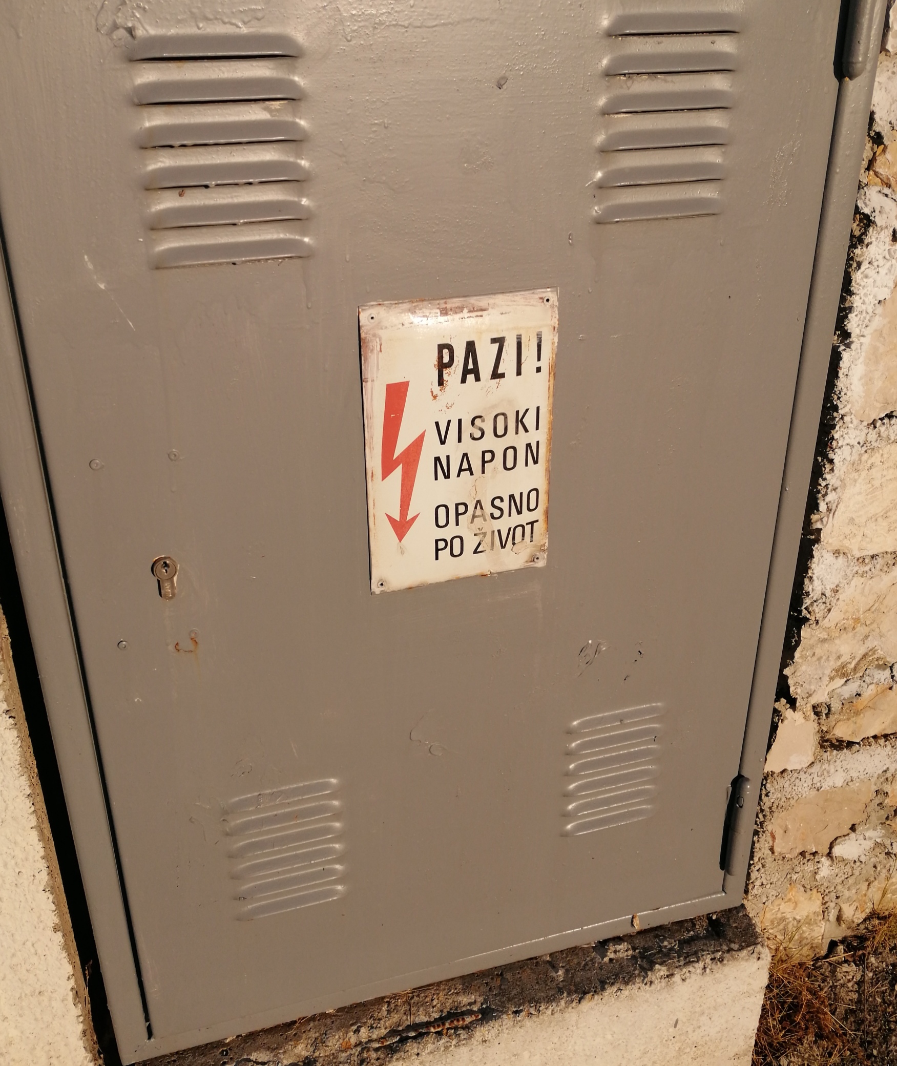 Szara skrzynka elektryczna na betonowej podstawie z tabliczką ostrzegawczą 'PAZI! VISOKI NAPON OPASNO PO ZIVOT' (Uwaga! Wysokie napięcie. Niebezpieczne dla życia) z symbolem błyskawicy.