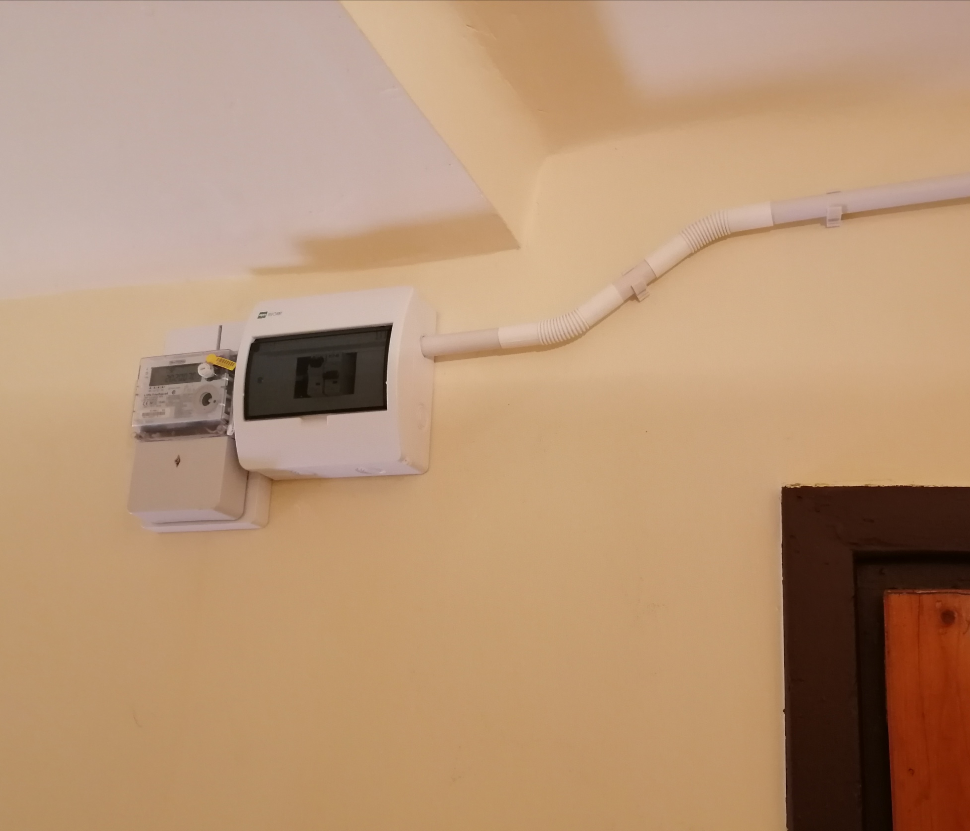 Zamontowany licznik elektryczny z białą skrzynką bezpiecznikową i peszlem na beżowej ścianie, widoczny fragment brązowej futryny drzwi.
