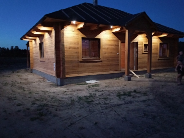 Drewniany dom jednorodzinny oświetlony zewnętrznymi lampami punktowymi pod okapem dachu, widok wieczorny z ziemistym podłożem.