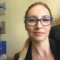 Portret kobiety w okularach, w biurowym otoczeniu z dokumentami i obrazami na ścianie, uśmiechnięta, w czarnej marynarce.