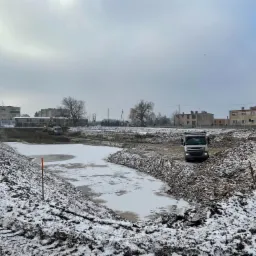 Zimowy krajobraz z wykopanym, częściowo zamarzniętym zbiornikiem wodnym. W tle budynki i maszyny budowlane, na pierwszym planie hałda ziemi i ciężarówka. Ślady opon na śniegu sugerują niedawne...