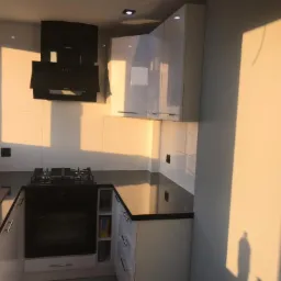 Nowoczesna kuchnia z białymi szafkami, czarnym blatem i płytą grzewczą, oświetlona popołudniowym słońcem, z widocznym okapem kuchennym.