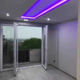 Pokój z szarymi ścianami, podłogą imitującą drewno i otwartymi, białymi drzwiami balkonowymi. Sufit podwieszany z oświetleniem LED w kolorze fioletowym.