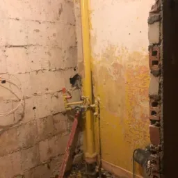 Stan surowy łazienki w Sandomierzu: żółta rura gazowa, ślady po starych płytkach na ścianie, gruz i poziomica w narożniku, przygotowania do prac wykończeniowych.