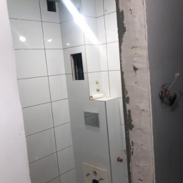 Michał Baska - Wnętrze łazienki w trakcie prac wykończeniowych, widoczne białe płytki na ścianach, częściowo zamontowana toaleta i sufit z płyt gipsowo-kartonowych, surowy ościeżnica drzwi z widocznymi przewodami...