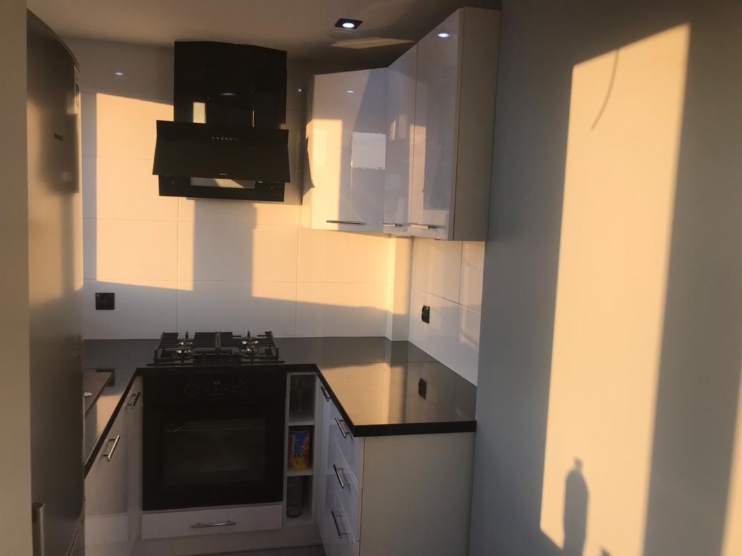 Nowoczesna kuchnia z białymi szafkami, czarnym blatem i płytą grzewczą, oświetlona popołudniowym słońcem, z widocznym okapem kuchennym.