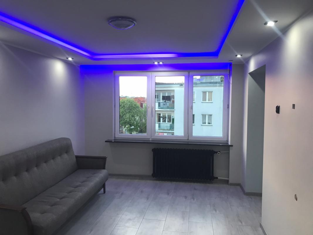 Jasne wnętrze po remoncie z szarą sofą, grzejnikiem pod oknem i podświetleniem LED sufitu na niebiesko.