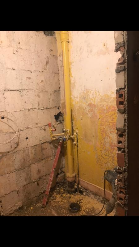 Stan surowy łazienki w Sandomierzu: żółta rura gazowa, ślady po starych płytkach na ścianie, gruz i poziomica w narożniku, przygotowania do prac wykończeniowych.