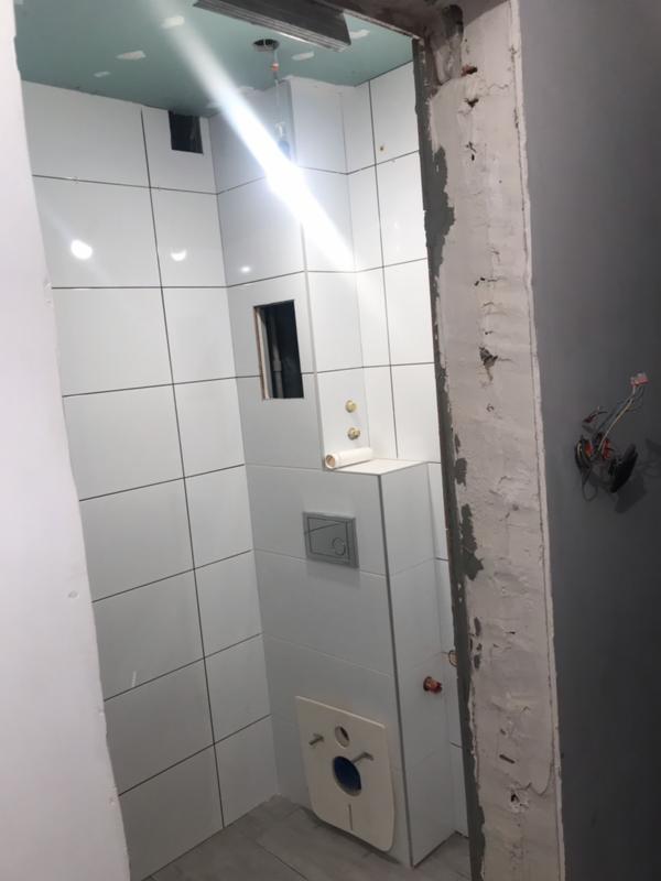 Wnętrze łazienki w trakcie prac wykończeniowych, widoczne białe płytki na ścianach, częściowo zamontowana toaleta i sufit z płyt gipsowo-kartonowych, surowy ościeżnica drzwi z widocznymi przewodami...