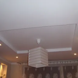 Biały sufit podwieszany z wbudowanymi punktowymi lampami LED i kwadratową wnęką, widoczny fragment pokoju z zasłonami i lampą wiszącą.