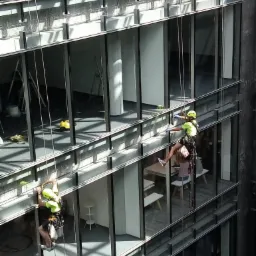 Dwaj alpiniści przemysłowi, zabezpieczeni linami, czyszczą szklaną fasadę nowoczesnego biurowca z widocznymi wnętrzami biur.