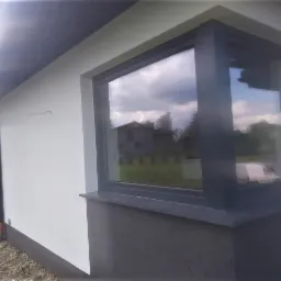 Fragment ściany zewnętrznej domu z narożnym oknem w grafitowej ramie, widoczne wykończenie elewacji w jasnym kolorze i cokół w ciemniejszym odcieniu, w oknie odbija się krajobraz z budynkiem.