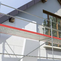 Fragment elewacji domu w trakcie ocieplania, widoczne rusztowanie, białe płyty izolacyjne, okno z żaluzjami i fragment dachu.