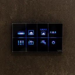 Szklany panel dotykowy z podświetlanymi ikonami sterowania funkcjami domu, zamontowany na szarej ścianie o porowatej strukturze, z widocznym logo IUVO w prawym dolnym rogu.