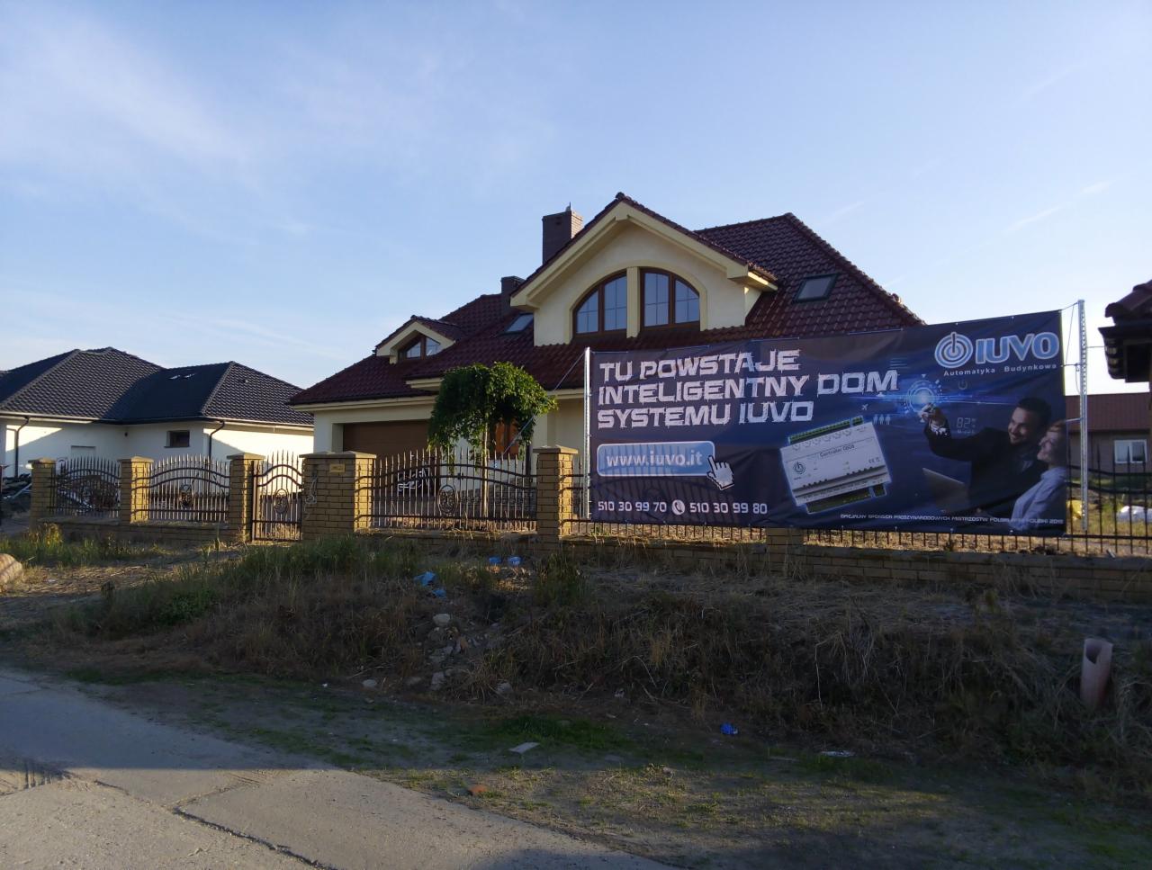 Baner reklamowy systemu inteligentnego domu IUVO zawieszony na ogrodzeniu posesji z domem jednorodzinnym w tle.