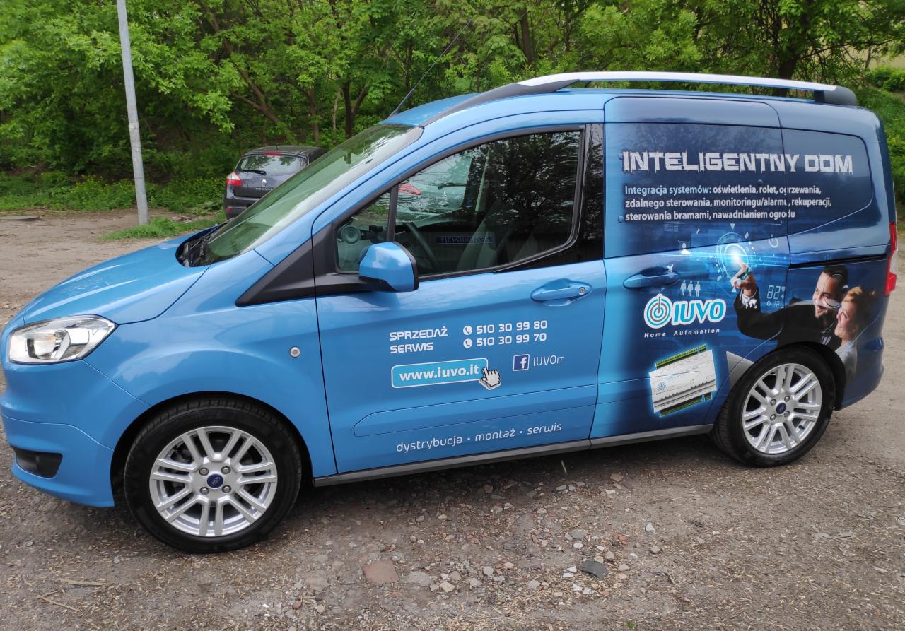 Niebieski samochód dostawczy z reklamą firmy oferującej rozwiązania dla inteligentnego domu, widoczne logo i dane kontaktowe firmy.