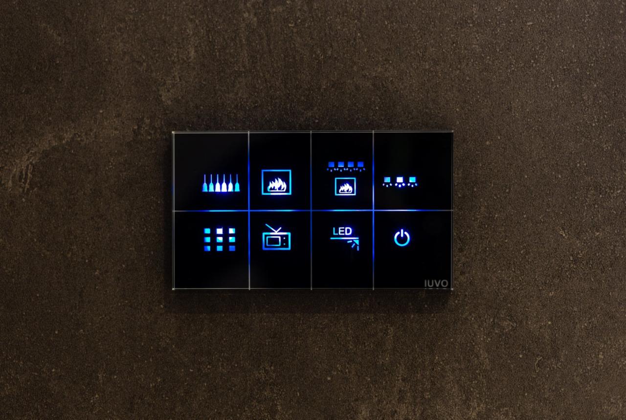 Szklany panel dotykowy z podświetlanymi ikonami sterowania funkcjami domu, zamontowany na szarej ścianie o porowatej strukturze, z widocznym logo IUVO w prawym dolnym rogu.