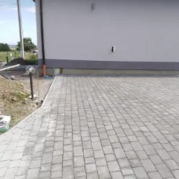 Ułożona szara kostka brukowa przed szarym budynkiem z widoczną rurą spustową, lampą ogrodową i fragmentem przygotowanego terenu.