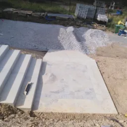 Budowa kamiennych schod&oacute;w i tarasu z kostki na gruncie, widoczne materiały budowlane, piasek i narzędzia brukarskie.