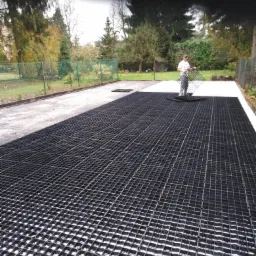 Montaż plastikowych kratek trawnikowych na przygotowanym podłożu, widoczny pracownik z kratką w ręku, w tle ogrodzenie posesji i drzewa.