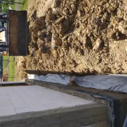 Wykop fundamentowy zabezpieczony folią kubełkową i geowłókniną, widoczna rura kanalizacyjna, ziemia z wykopu oraz koparko-ładowarka w tle.