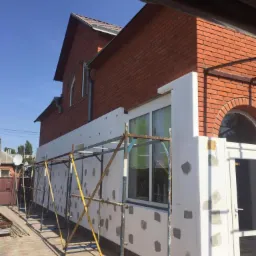Dom z czerwonej cegły w trakcie ocieplania elewacji, widoczne rusztowania i płyty styropianowe z zaprawą klejową.
