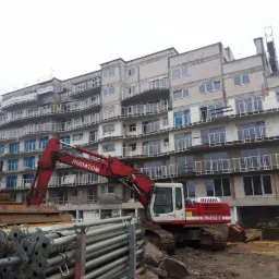 Wielopiętrowy budynek w trakcie budowy z widoczną czerwoną koparką firmy RUDATOM na pierwszym planie, stosy szalunków i materiałów budowlanych, pochmurne niebo.