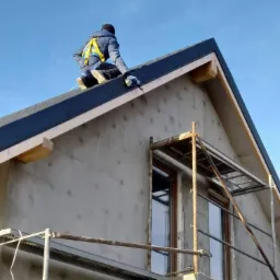 Pracownik w uprzęży zabezpieczającej montuje obróbkę blacharską na krawędzi dachu nowo wybudowanego domu ze ścianami w surowym tynku, widoczne rusztowanie.